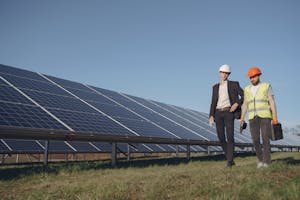 Full body of young focused manager in formal wear and electrical technician in uniform walking on path near solar panels and discussing working process